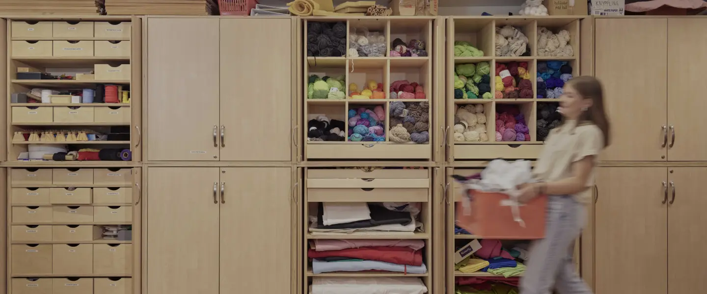 En tjej bär på en låda med tyg. Skåp med garn och tyger i bakgrunden